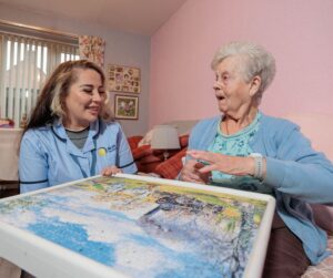 Image of a Care Assistant and an elderly lady, sat on a couch whilst playing a jigsaw puzzle.