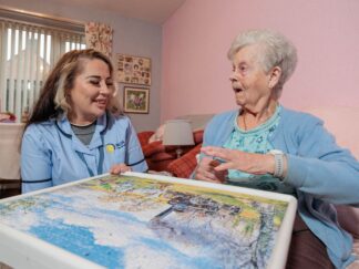 Image of a Care Assistant and an elderly lady, sat on a couch whilst playing a jigsaw puzzle, highlighting our companionship services.