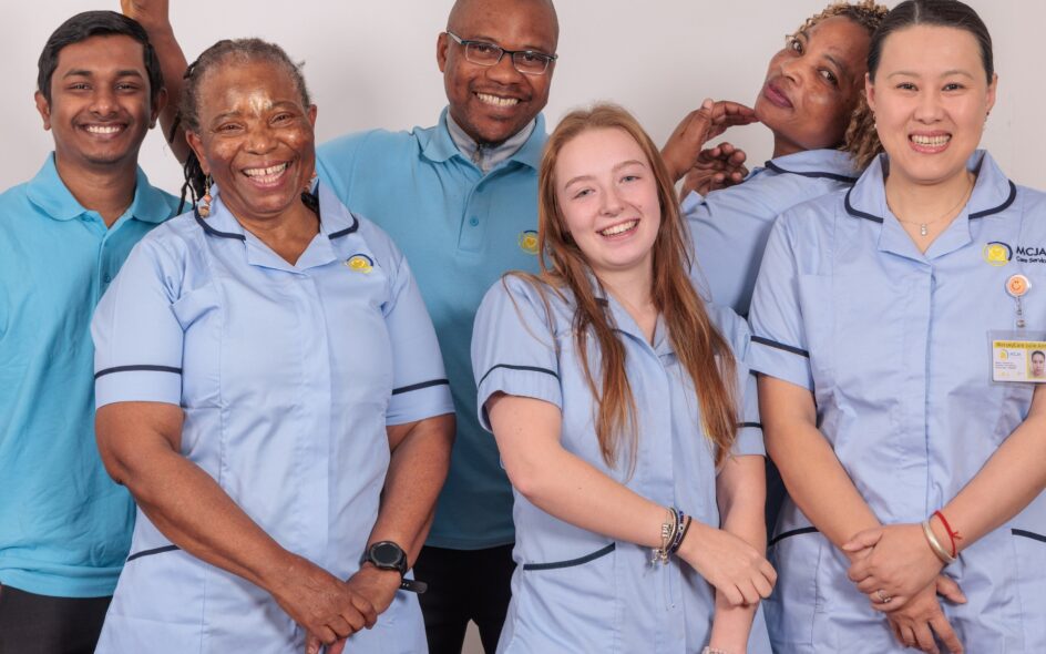 A group of 5 Care Assistants wearing a blue uniform smiling and posing for the camera, in front of a white wall.