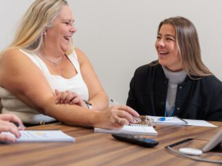 2 women sat in a meeting, smiling and laughing.