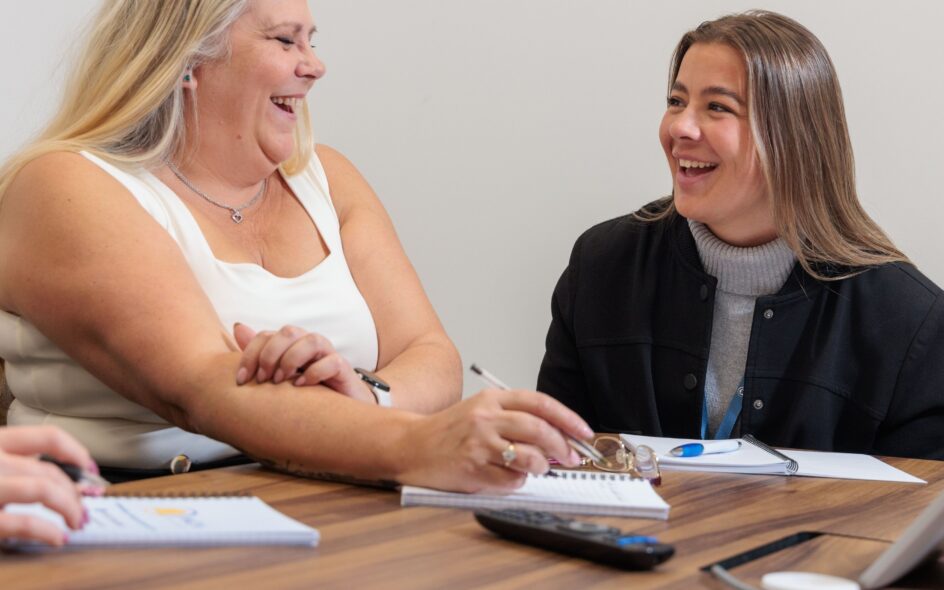 2 women sat in a meeting in an office setting, smiling and laughing.