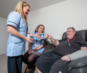 Image of 2 Care Assistants in a blue uniform and an man in a home setting, passing over a cup of tea.