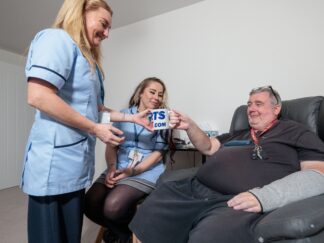 Image of 2 Care Assistants in a blue uniform and an man in a home setting, passing over a cup of tea. voices of care