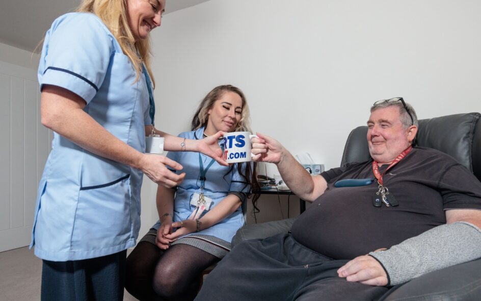 Image of 2 Care Assistants in a blue uniform and an man in a home setting, passing over a cup of tea. voices of care