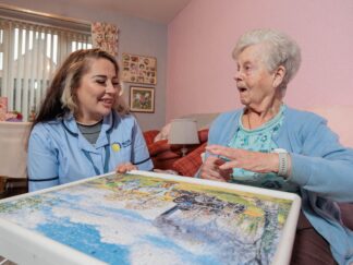 Image of a Care Assistant in a blue uniform and an elderly lady, sat on a couch whilst playing a jigsaw puzzle.