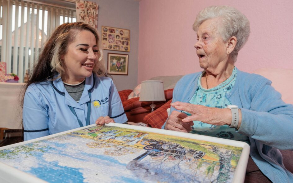 Image of a Care Assistant in a blue uniform and an elderly lady, sat on a couch whilst playing a jigsaw puzzle. listening in care