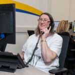 An image of a woman at a desk in an office environment, answering a phone call.