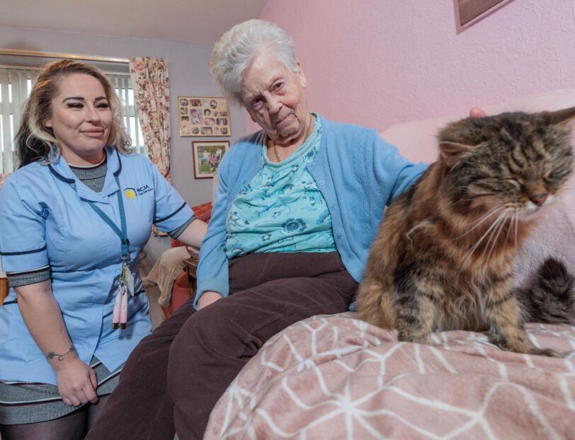 Image of a Care Assistant and an elderly lady, sat on a couch whilst petting a cat.