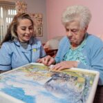 Image of a Care Assistant and an elderly lady, sat on a couch whilst playing a jigsaw puzzle.