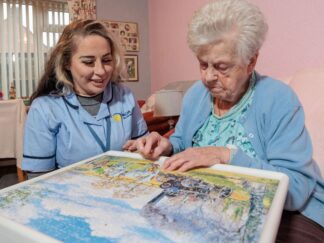 Image of a Care Assistant and an elderly lady, sat on a couch whilst playing a jigsaw puzzle.