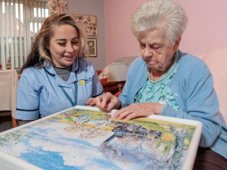 Image of a Care Assistant and an elderly lady, sat on a couch whilst playing a jigsaw puzzle.