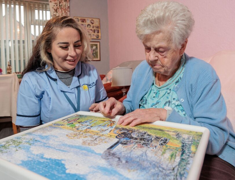 Image of a Care Assistant and an elderly lady, sat on a couch whilst playing a jigsaw puzzle.