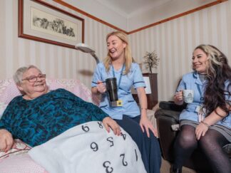 Image of 2 Care Assistants and an elderly lady in a home setting, passing over a cup of tea.