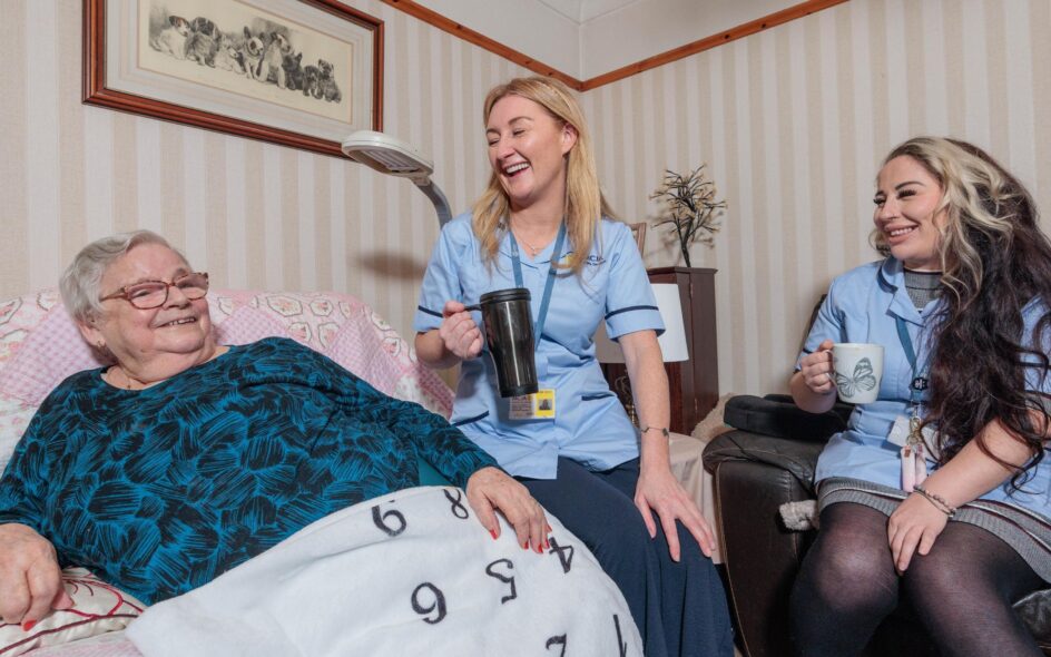 Image of 2 Care Assistants and an elderly lady in a home setting, passing over a cup of tea.