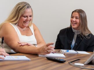 2 women sat in a meeting, smiling and laughing.