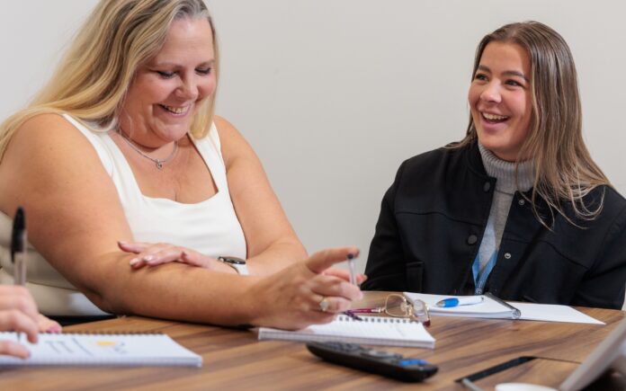 2 women sat in a meeting, smiling and laughing.