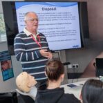 An image of a man stood in a classroom, teaching a group of students and presenting a powerpoint.