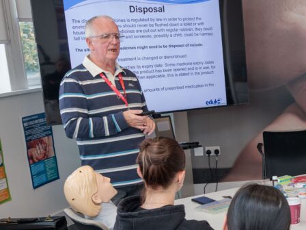 An image of a man stood in a classroom, teaching a group of students and presenting a powerpoint.
