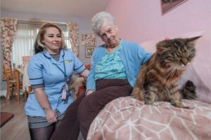 An image of a care assistant in a blue uniform and a service user in a blue top and blue cardigan, petting a cat whilst sat on a pink couch.