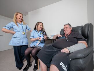 Two care assistants in a blue uniform, handing over a cup of tea to a service user in a care home setting.