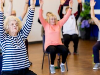 An image of a group of elderly people taking part in chair yoga in a gymnasium hall.