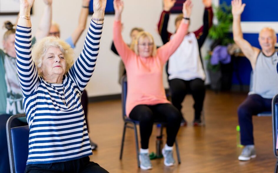 An image of a group of elderly people taking part in chair yoga in a gymnasium hall.