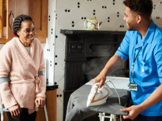 An image of a male carer in a blue uniform in a home setting providing care to a disabled woman, whilst completing ironing duties.