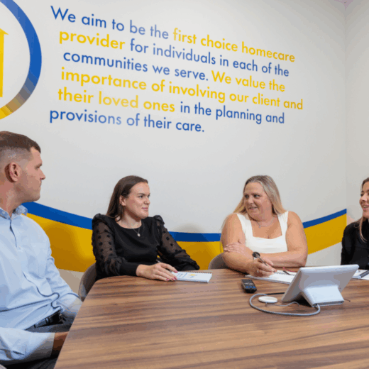 A man and 3 women in an office setting taking part in a meeting, laughing and smiling at each other.