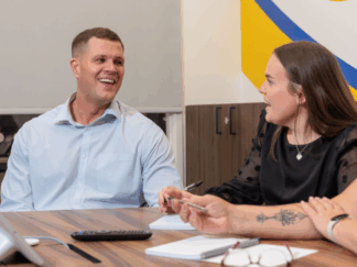 A man and a woman in an office setting taking part in a meeting, laughing and smiling at each other.