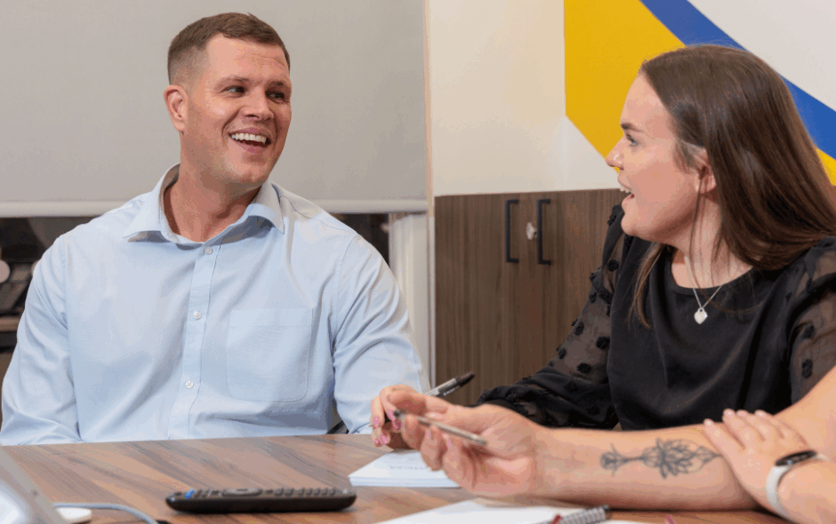 A man and a woman in an office setting taking part in a meeting, laughing and smiling at each other.